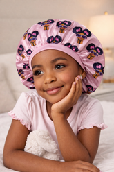 Child wearing a pink satin bonnet with cartoon character design, lying on a bed.