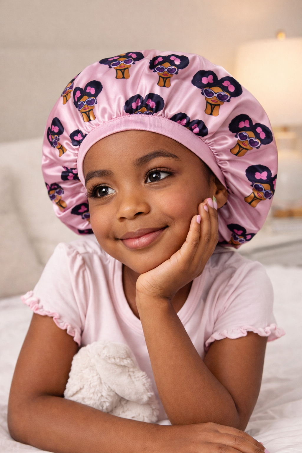 Child wearing a pink satin bonnet with cartoon character design, lying on a bed.