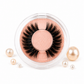 False eyelashes in a clear container with a pinkish-beige background, surrounded by pearls on a white background.