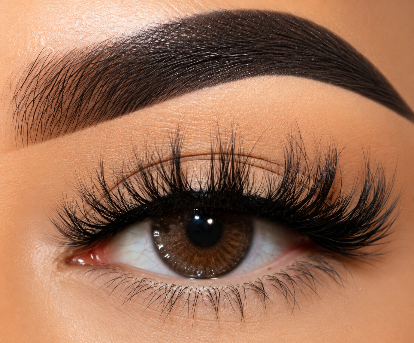 Extreme close-up of a human eye with long, dark false eyelashes applied, a defined dark brown eyebrow, and warm beige skin.