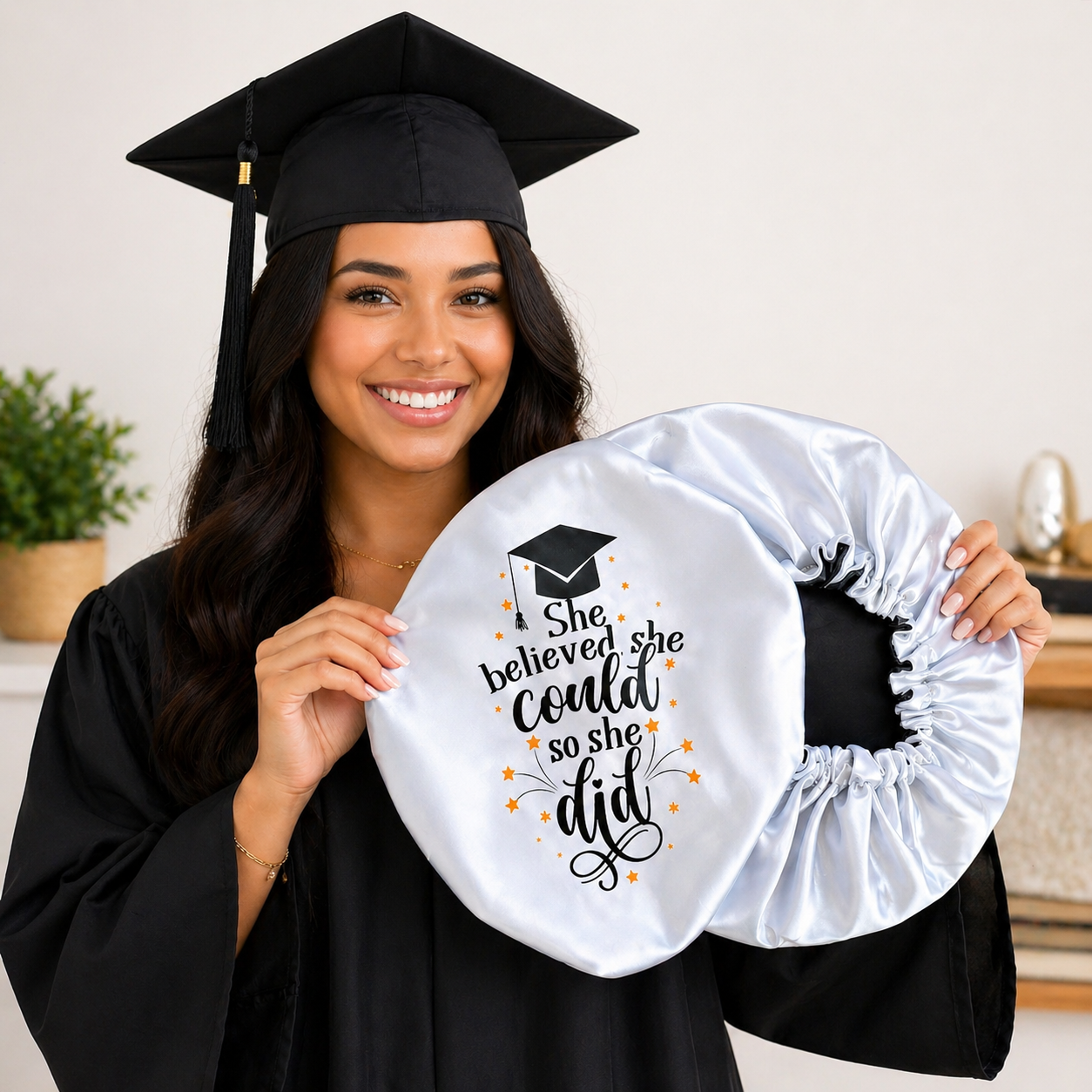 Graduate holding a white satin circle with text, wearing a black graduation cap and gown.