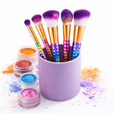 Makeup brushes with colorful bristles in a purple holder, surrounded by glittery makeup products on a white background.