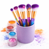 Makeup brushes with colorful bristles in a purple holder, surrounded by glittery makeup products on a white background.