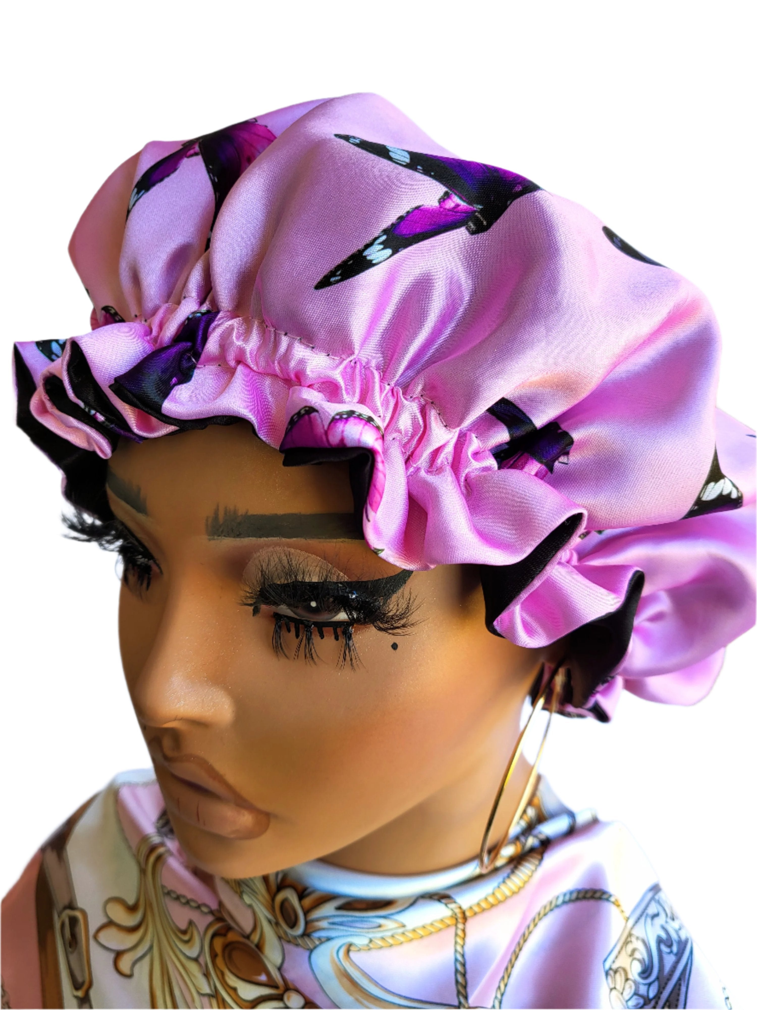 A woman wearing a pink satin bonnet with a butterfly pattern. The bonnet is tied around the head, and the woman is posed with her eyes closed, showcasing the product&